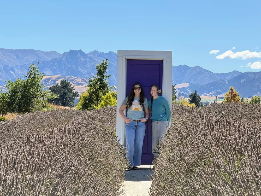 Emily and Mia standing in a freestanding purple door set in the middle of a lavender field, mountains across the valley behind