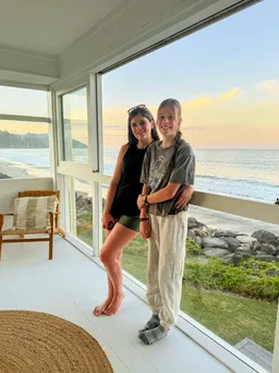 Emily and Mia together on the white veranda of the beach house, the sea and a pale sunset filling the open window behind them
