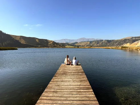 Mia and Case sitting together at the end of a long wooden jetty, looking out over the lake to the hills beyond