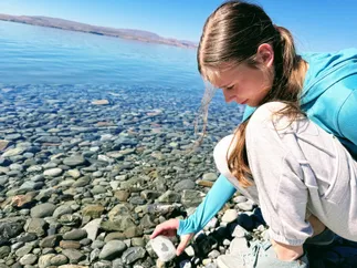 Mia crouching at the water's edge reaching for a pale stone marked with a heart, clear shallows and rounded pebbles all around