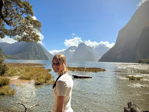 Mia turning to the camera at the water's edge at Milford Sound, the fjord walls and peaks rising into cloud behind her