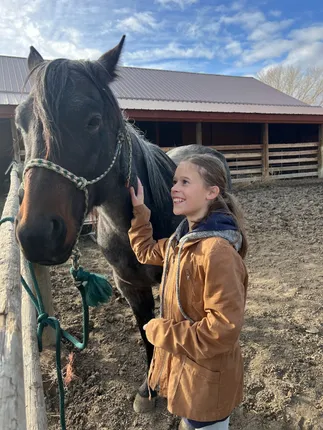 Mia in a canvas coat laughing up at a dark bay horse tied at the rail, the barn behind them