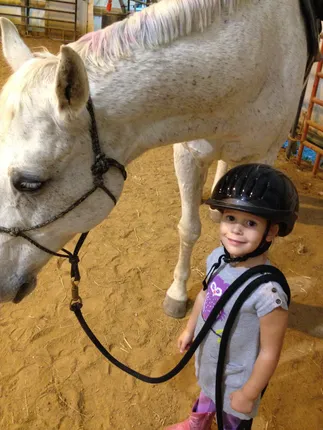 Mia as a toddler in a riding helmet holding the lead rope of a big grey pony in an indoor arena
