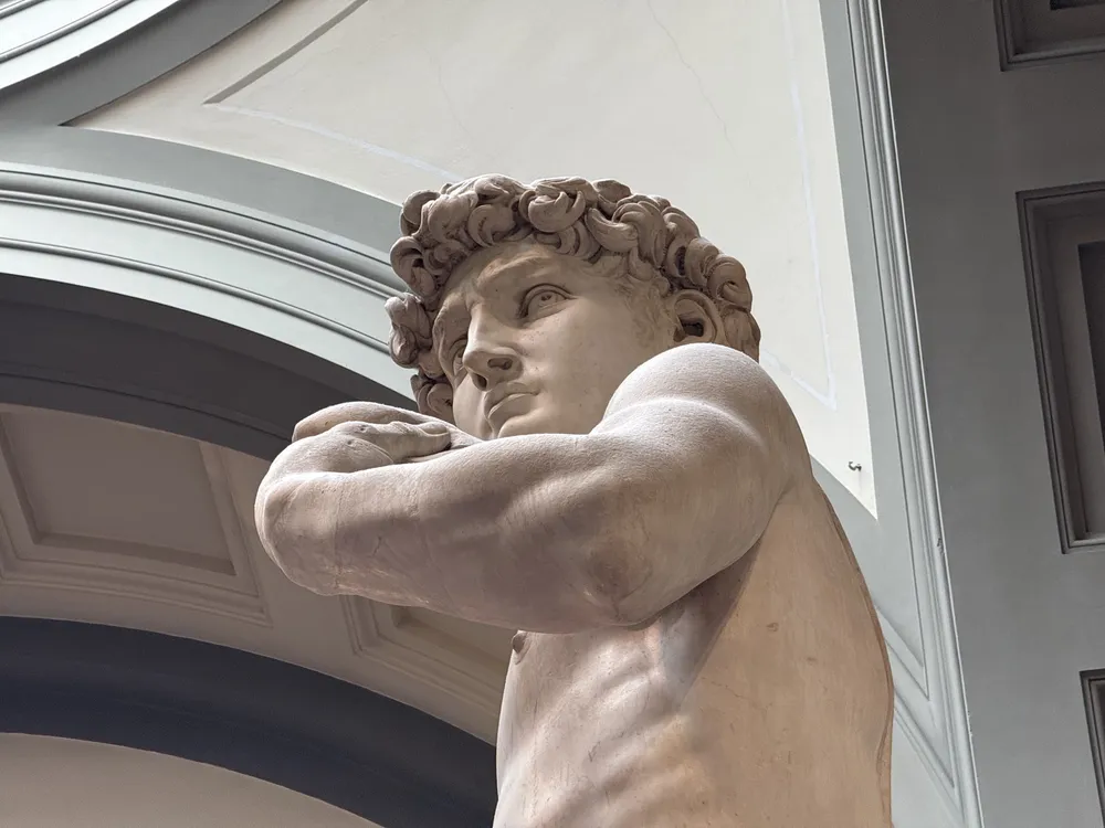 Michelangelo's David from below, the head turned and the raised arm resting on the shoulder, under the gallery's vaulted ceiling