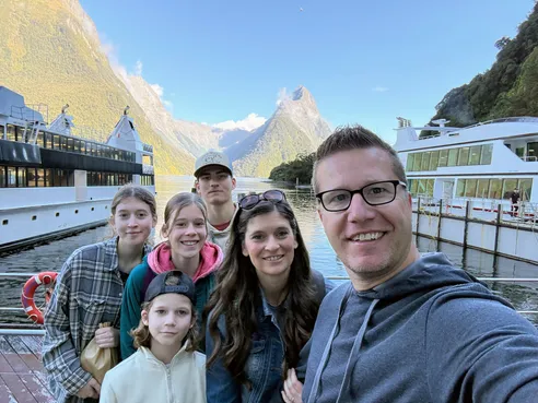 A family selfie on the wharf at Milford Sound, all six of them with Mitre Peak and the cruise boats behind