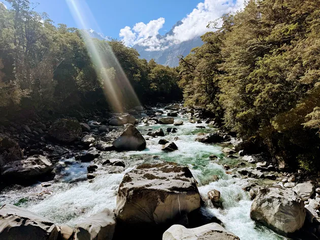 A clear green river running over boulders through native bush, sunbeams slanting down and a peak in cloud at the head of the valley