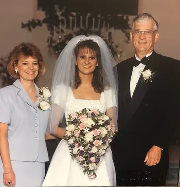 Christy in her wedding gown holding a cascade bouquet, flanked by her mother and father
