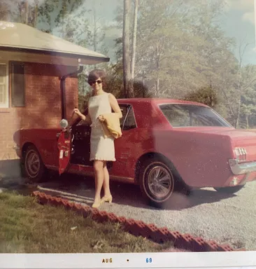 A 1969 snapshot of Emily's mother in a pale shift dress and sunglasses beside a red Mustang on the drive