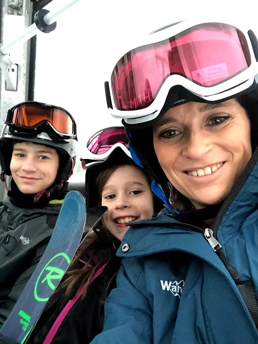 Emily grinning in ski goggles on the chairlift with two of the children beside her