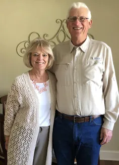 Emily's parents standing together indoors, she in a cream crocheted cardigan and pink flower necklace