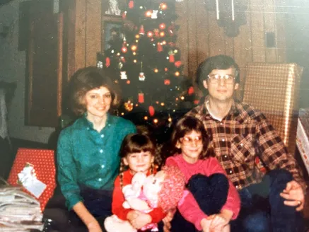 The four of them in front of a lit Christmas tree, the two girls on the floor with a doll and a toy heart