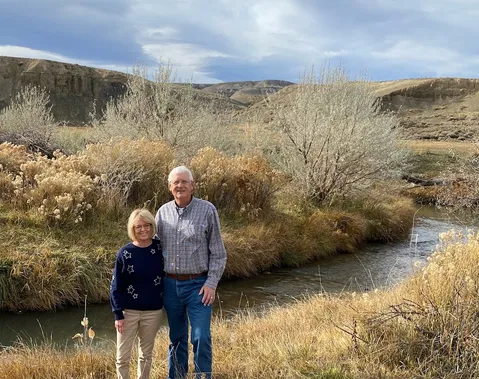 Emily's parents standing by the creek at the ranch, sage and autumn grass around them and the bluffs behind
