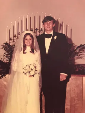 Emily's parents on their wedding day, the bride in a high-necked lace gown and long veil, the groom in a black tuxedo, candelabra behind them