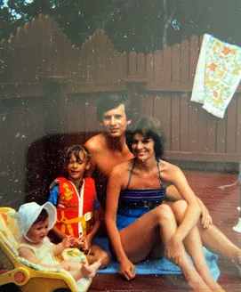 The young family on a deck by the pool, the parents in swimsuits with a small girl in a life jacket and a baby in a bouncer