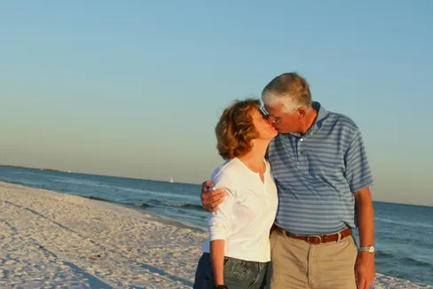 Emily's parents kissing on a white sand beach at golden hour, the sea and a distant sail behind them
