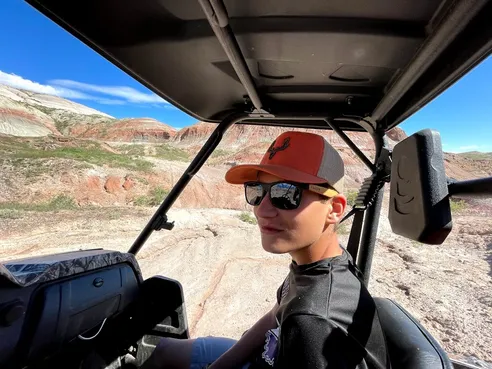 Preston at the wheel of a side by side in a cap and sunglasses, banded red and white badlands behind him
