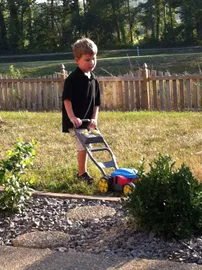 Preston in a black polo pushing a toy lawnmower across the grass, a picket fence behind him