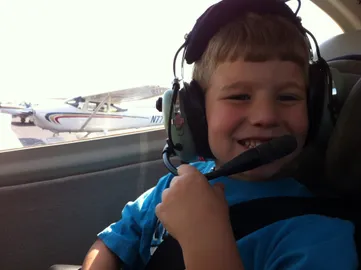Preston in the back of a small aeroplane in an oversized headset, grinning, another Cessna out the window