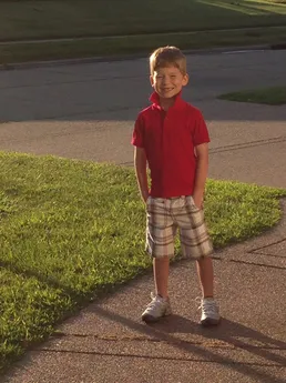 Preston in a red polo and plaid shorts standing on the drive in low evening sun, hands in his pockets