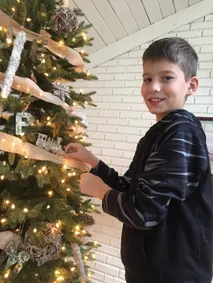 Preston hanging a silver train ornament on the lit Christmas tree, turning to smile at the camera