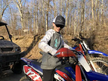 Preston in a cap and sunglasses standing over a blue and red dirt bike in bare winter woods