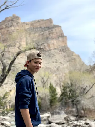 Preston in a backwards cap and navy hoodie by a creek, a tall limestone bluff rising behind him