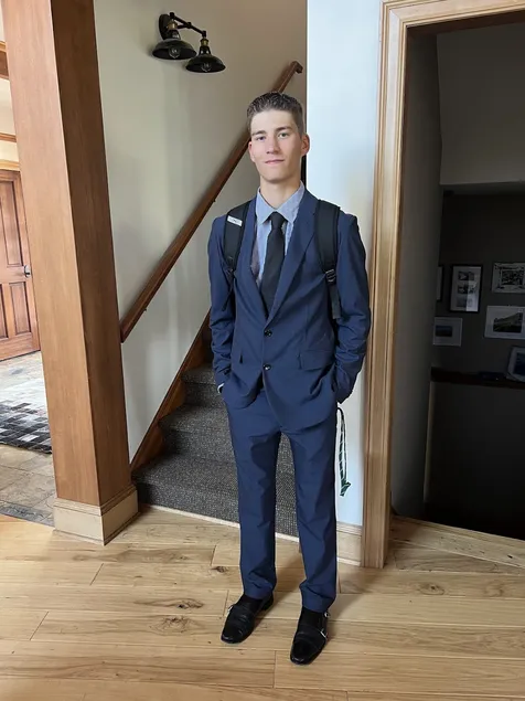 Preston in a navy three piece suit and tie with a backpack on, standing in the hall by the stairs