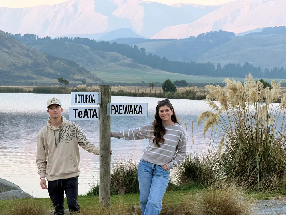 Preston and Emily either side of a wooden signpost reading Hoturoa, Te Ata and Paewaka, a lake and pink lit mountains behind them