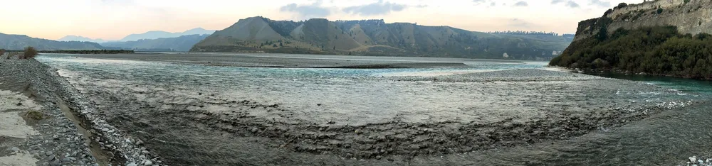 A wide panorama of a braided river running over grey shingle flats at dusk, bluffs on the right and hills fading into haze