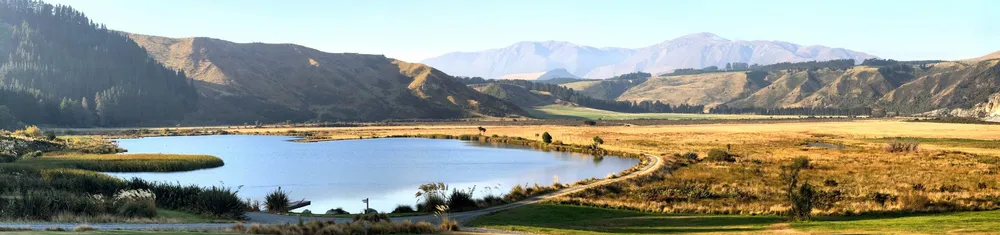 A wide panorama of a still lake below golden hills and distant ranges, a gravel track curving away along the shore