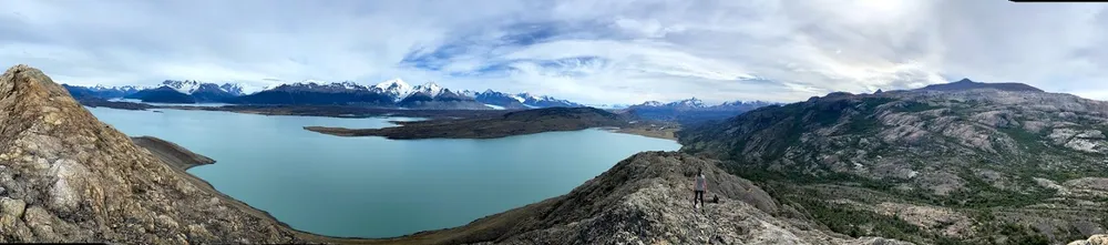 A person standing on a rocky peak overlooking a wide vista of turquoise glacier lakes and snow mountains