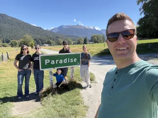 Josh taking a selfie with the whole family behind him at the green Paradise road sign, mountains and farmland beyond