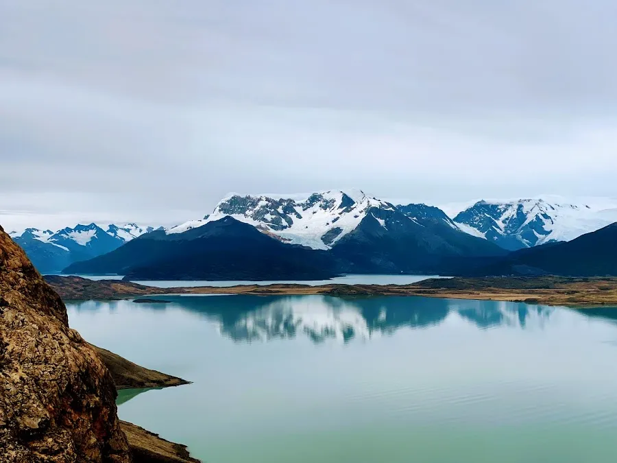 A still turquoise lake reflecting a long line of snow-covered peaks under flat grey cloud