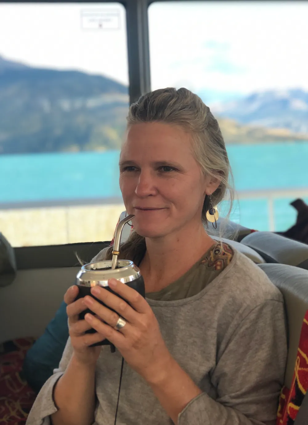 A woman on the bus holding a mate gourd with its metal straw, the turquoise lake through the window behind her