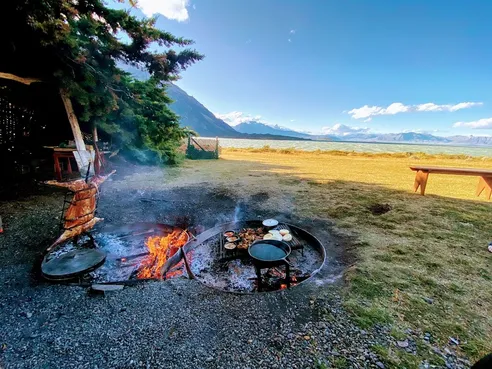 A fire pit with cast iron cooking gear roasting lamb, snow-capped peaks across the valley