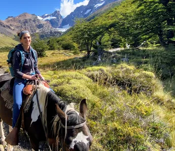 A woman on horseback on a mountain trail, a glacier and rock spires in the valley behind her