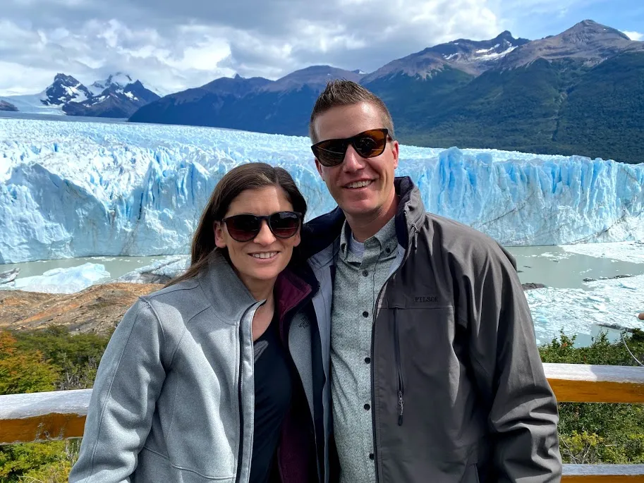 Emily and Josh together in sunglasses and jackets, the Perito Moreno glacier and mountains filling the view behind them