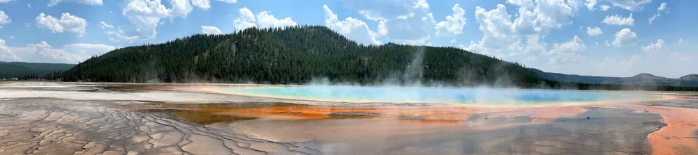 The Grand Prismatic Spring, steam lifting off bands of orange and turquoise, dark forest on the ridge behind