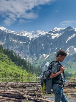 Preston with a backpack walking the driftwood shore of Avalanche Lake, waterfalls streaking the snowy cirque behind