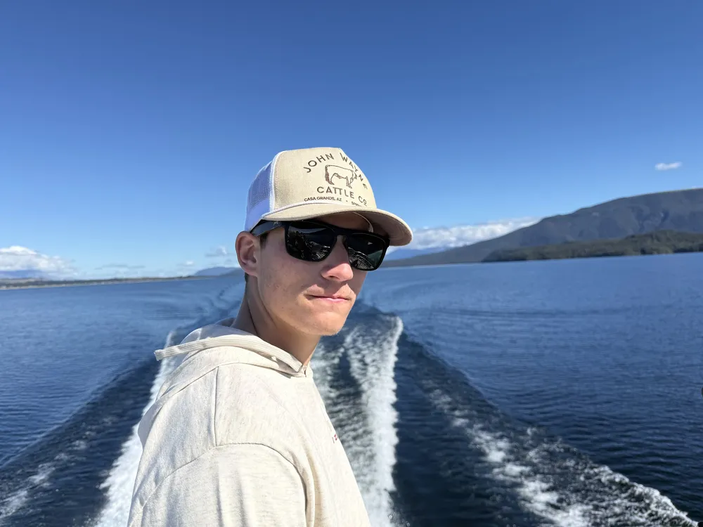 Preston in a cap and sunglasses looking back from the stern of a boat, the wake running away across the lake behind him