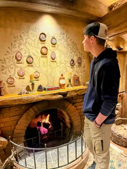Preston standing at the hearth inside Bag End looking up at the painted family tree of portraits above the mantel, a fire lit below