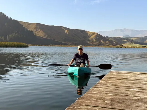 Preston paddling a turquoise kayak towards the jetty in a cap and sunglasses, green hills rising behind the lake