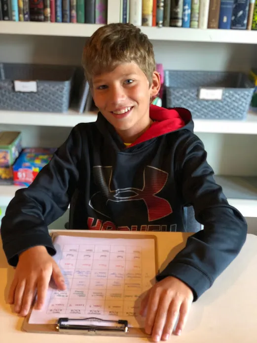 Preston grinning at the schoolroom table with his weekly assignment sheet on a clipboard in front of him, bookshelves behind