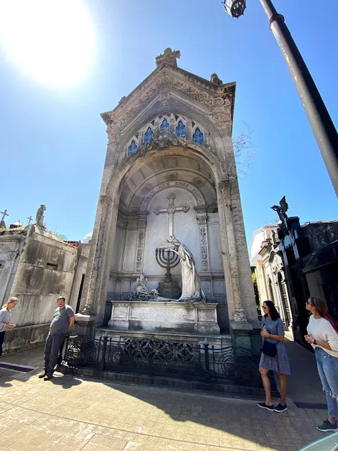 A tall ornate mausoleum with a cross at its apex, visitors walking past it in the Recoleta Cemetery