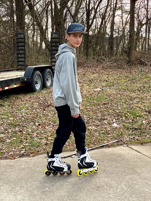 Preston in a cap and hoodie standing on inline skates on the drive, bare winter woods behind