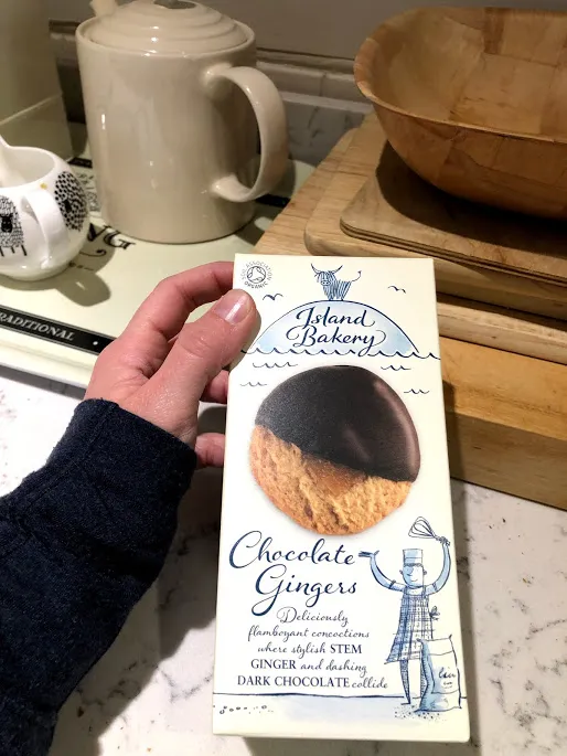 A hand holding a box of Island Bakery Chocolate Gingers in a kitchen, kettle and wooden bowls behind