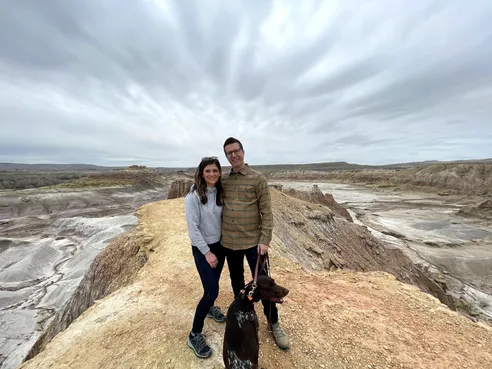 Emily and Josh with the dog on a ridge of banded grey and yellow badlands at Devils Kitchen under heavy cloud