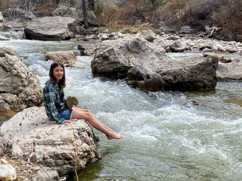 Evie in a checked shirt sitting on a limestone slab with her bare feet over the creek, white water running past