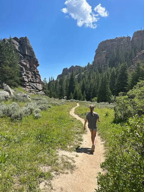 Mia walking away up a narrow dirt path through sage and meadow towards granite outcrops and firs