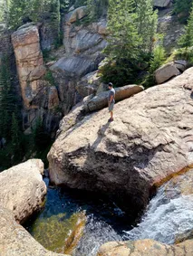 Josh standing on a great sloping boulder above a deep green plunge pool, the canyon walls and firs behind